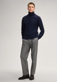 Junger Mann in einem dunkelblauen, kabelgestrickten Rollkragenpullover, grauen, plissee Hosen und dunkelbraunen Wildlederstiefeln, der mit den Händen in den Taschen steht.