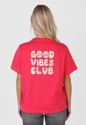 Person med langt blondt hår iført en rød T-shirt med teksten "GOOD VIBES CLUB" og smileyer på ryggen, stående mod en ensfarvet baggrund.