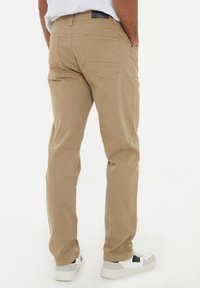 Beige Baumwollhose mit geradem Schnitt, zwei Gesäßtaschen und einem Lederpatch am Bund. Getragen mit weißen Sneakers.