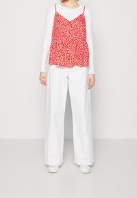 Top sin mangas rojo con lunares blancos, superpuesto sobre una camisa blanca de mangas largas; combinado con pantalones anchos blancos y zapatillas blancas.