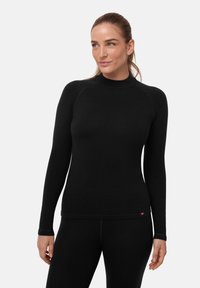 Frau mit hellbraunen, zurückgebundenen Haaren, die ein eng anliegendes schwarzes Langarmshirt und schwarze Hose trägt, vor einem schlichten weißen Hintergrund stehend.