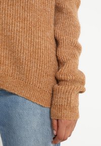 Strickpullover in Hellbraun, mit gerippten Ärmeln und Saum. Weiche Textur mit sichtbaren Nahtdetails.