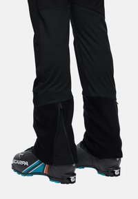 Zwarte skibroeken met ritssluitingen bij de enkels, gecombineerd met zwarte skibotten met blauwe en oranje accenten.