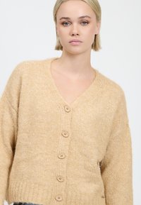 Beige gestrickte Strickjacke mit V-Ausschnitt, ausgestattet mit fünf Holzknöpfen auf der Vorderseite und einer strukturierten, weichen Optik.