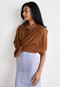 Bruine geknoopte blouse met een zachte textuur, gecombineerd met een lichtblauwe kokerrok met verticale strepen in meerdere kleuren.