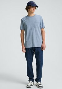 Tricou scurt de culoare albastru deschis, cu buzunar pe piept, asortat cu blugi de culoare albastră închisă și adidași alb-negru. Șapca este de culoare bleumarin, cu litere albe.