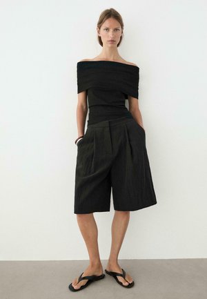 Kvinde iført sort off-shoulder top, løse sorte knælange shorts, sorte sandaler, står op ad hvid væg med hænderne i lommerne.