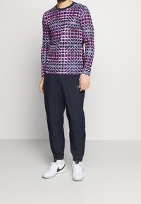 Man i en långärmad skjorta med geometrisk mönstring, mörkblå joggarbyxor, vita strumpor och vita sneakers från Nike med svart swoosh.