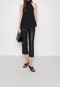 Svart ärmlös halterneck-topp, lös passform. Glansiga svarta lädercroppade byxor. Svarta klacksandaler med knytt design. Håller en svart clutch.