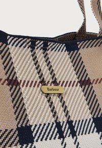 Τσάντα ώμου από καρό ύφασμα με μπεζ, μαύρα και καφέ τετράγωνα και μικρή μεταλλική χρυσή ετικέτα με την επιγραφή "Barbour" κοντά στην κορυφή.