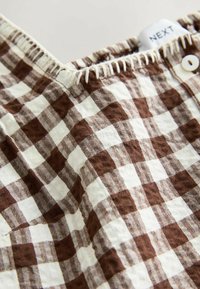 Tissu à carreaux marron et blanc avec bouton et ceinture élastique cousue, montrant l'étiquette de la marque "NEXT" sur un vêtement.