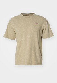 Camiseta de manga corta, color marrón claro, fabricada en un tejido suave y texturizado. Presenta un pequeño parche de logo en el pecho y un clásico escote redondo.