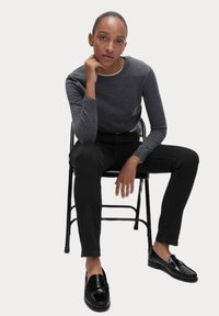 Graues Langarmshirt mit rundem Ausschnitt, schwarze eng anliegende Hose und schwarze Slipper an einer sitzenden Figur vor einem schlichten Hintergrund.