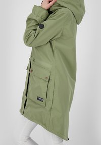 Parka lunga verde realizzata in materiale impermeabile, con cappuccio, numerose tasche e una patch nera con logo sul davanti.