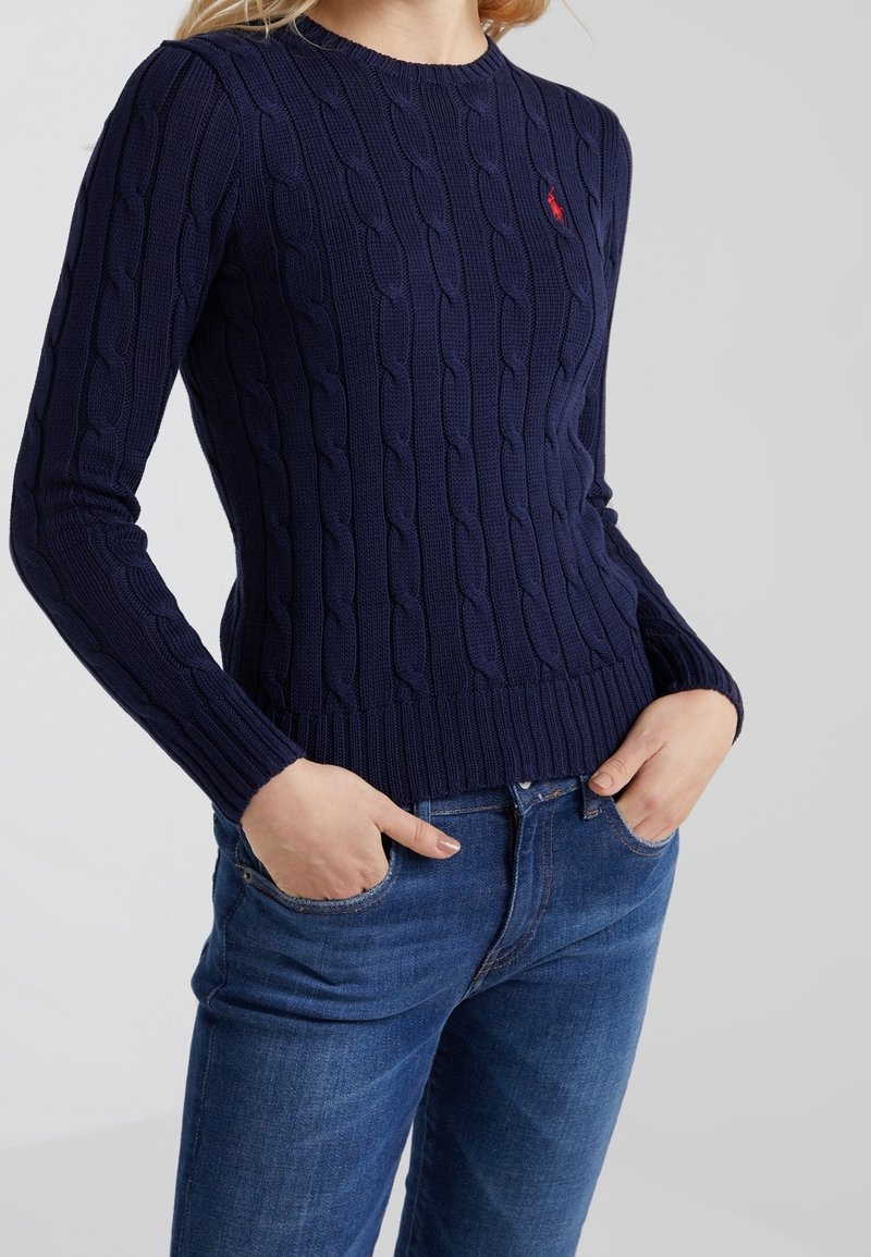 Marineblauer, kabelgestrickter Pullover mit langen Ärmeln, Rundhalsausschnitt und gerippten Bündchen. Verfügt über ein kleines rotes Logo auf der Brust. Kombiniert mit blauen Jeans.