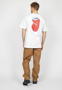 White t-shirt with a red soda can graphic on the back. Brown pants and black sneakers. Casual design with short sleeves.