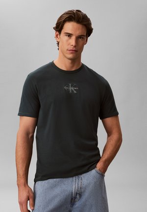 Junger Mann trägt ein schwarzes Calvin Klein T-Shirt und hellblaue Jeans, steht vor einem schlichten grauen Hintergrund.