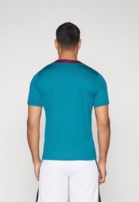 Teal lyhythihainen urheilupaita, jossa on violetti kaulus, hengittävä rakenne ja istuva muotoilu. Takakuva näyttää paidan rakenteen.