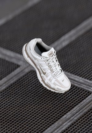 Weißer und beiger Nike Laufschuh mit Schnürsenkeln liegt auf einer dunklen Metallgitternfläche, dargestellt aus einem schrägen Blick von oben.