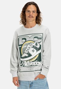 Man met krullend haar en een snor, gekleed in een grijze sweatshirt met een grote afbeelding van een sneaker en het woord "Athletics."
