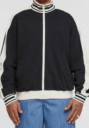 Person trägt eine schwarz-weiße Reißverschlussjacke mit gestreiftem Kragen und Bündchen, kombiniert mit blauen Jeans, Hände entspannt an den Seiten.