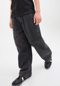 Crne traperice opuštenog kroja, s ravnim nogavicama, vidljivim šavovima i blago izblijedjelom bojom. Uparene s crnim tenisicama.