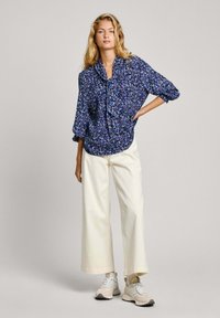 Blusa fluida blu navy con stampa floreale, pantaloni ampi color avorio abbinati e sneakers chiare. La blusa presenta un colletto a fiocco e maniche 3/4.