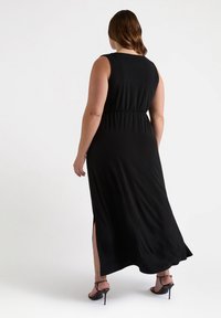 Abito maxi nero con design senza maniche, vita segnato e spacchi laterali. Tessuto dalla texture liscia. Indossato con sandali con tacco alto.