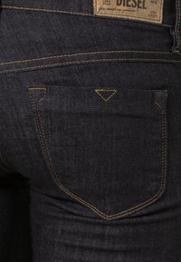 Ciemnoniebieskie dżinsy z kontrastującymi żółtymi szwami. Posiadają unikalny trójkątny detal szwu na tylnej kieszeni. Widoczna metka marki.