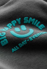 Šedá látka s vyvý šeným tyrkysovým textem: "VELKÝ ŠŤASTNÝ ÚSMĚV" a "CELÝ DEN - KAŽDÝ DEN", spolu s grafikou smajlíka.