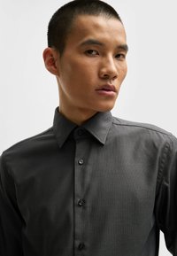 Chemise boutonnée gris foncé avec un motif texturé, col étalé et six boutons noirs, présentant des coutures discrètes le long des épaules.
