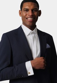 Navy suit jacket with a notched lapel, white dress shirt, and gold cufflinks. Pocket square features a blue checkered pattern.
