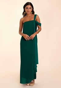 Grünes, einseitig getragenes Maxikleid aus fließendem Stoff, mit einem geknoteten Ärmeldetail und einem gestuften Saum, kombiniert mit klobigen goldenen Plateauschuhen.