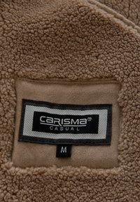 Brunt fleecetyg med en mjuk, texturerad yta och en påsydd svart etikett med texten "CARISMA" och "CASUAL", storlek "M".