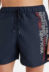 Bañador de marinero con cintura elástica y cordón, que presenta un logotipo impreso y texto en blanco y rojo en el lado izquierdo.