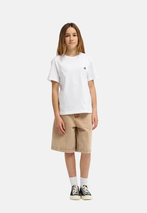 Jeune personne debout, portant un t-shirt blanc uni, un short beige jusqu'aux genoux, des chaussettes blanches et des baskets noires sur fond blanc.