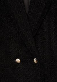 Gros plan d'un blazer noir texturé à double boutonnage avec des boutons décoratifs dorés et des revers crantés.