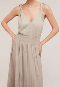 Robe longue beige clair avec décolleté en V, texture côtelée, bretelles croisées et taille élastique. Jupe plissée fluide.