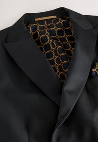 Black blazer featuring a satin lapel, with a geometric patterned lining in gold and navy. Includes a pocket square with matching design.
