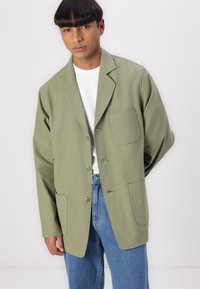 Olive green blazer with a relaxed fit, notch lapels, and two front pockets, paired with a white t-shirt and light blue jeans.