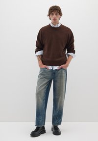 Brauner gerippter Strickpullover über einem weißen Kragenhemd, kombiniert mit hellblauen weit geschnittenen Jeans mit verwaschenen Stellen und schwarzen klobigen Schuhen.