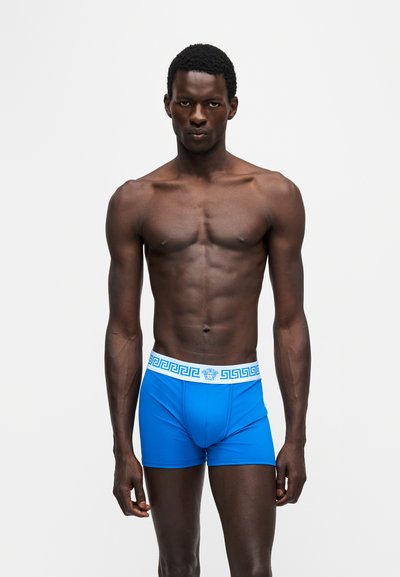 Mannequin masculin musclé à la peau foncée, debout face à l’avant, portant un boxer bleu vif avec une ceinture blanche et un motif clé grec bleu.