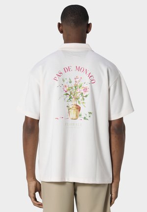 Mann trägt ein weißes Kurzarmhemd mit einem Blumenvasen-Design und dem Text „PAS DE MONACO“ auf dem Rücken.