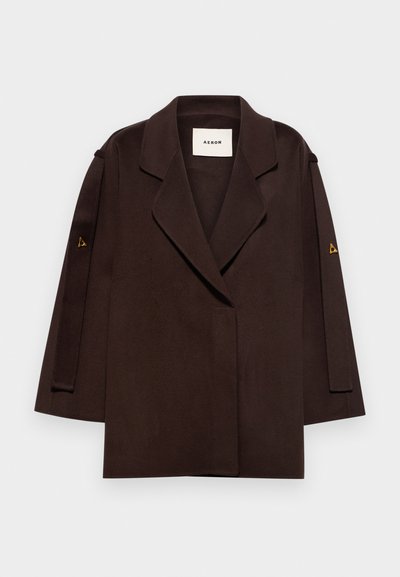 Hnědý přizpůsobený sako se strukturovaným límcem, širokými rukávy a trojúhelníkovým zlatým akcentem na manžetách. Hladká textilie, dvouřadý design.