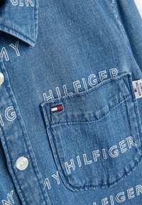 Jasnoniebieska koszula dżinsowa z białym haftowanym napisem. Klasyczny kołnierzyk, kieszeń na piersi z małą metką w kolorze czerwonym, białym i niebieskim. Zapięcie na guziki.