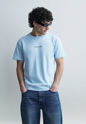 Jauns vīrietis ar cirtainiem matiem, valkājot melnus saulesbrilles, gaiši zilu T-kreklu "The Kooples" un zilas džinsas, stāvot ar rokām kabatās.