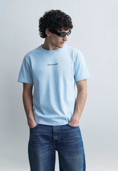 Jeune homme aux cheveux bouclés portant des lunettes de soleil noires, un t-shirt bleu clair "The Kooples" et un jean bleu, debout les mains dans les poches.