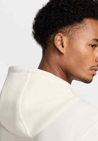 Felpa con cappuccio color crema dal texture morbido, cuciture visibili e vestibilità rilassata. Il profilo del modello è parzialmente visibile su uno sfondo chiaro.