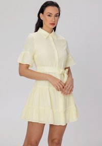 Robe jaune clair avec des manches à volants courtes, un devant à boutons et une ceinture à la taille. Présente un design de jupe à étages et un tissu texturé.