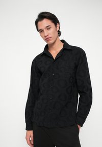 Fiatal férfi, sötét hajjal, fekete mintás hosszú ujjú ingben és fekete nadrágban, sima háttér előtt.